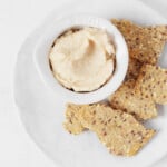 An overhead image of a small, pinch pot filled with a creamy white bean dip. The dip is served on a round white plate and accompanied by seeded crackers.