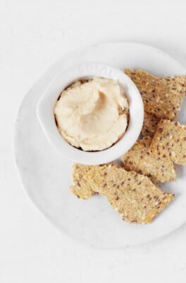 An overhead image of a small, pinch pot filled with a creamy white bean dip. The dip is served on a round white plate and accompanied by seeded crackers.