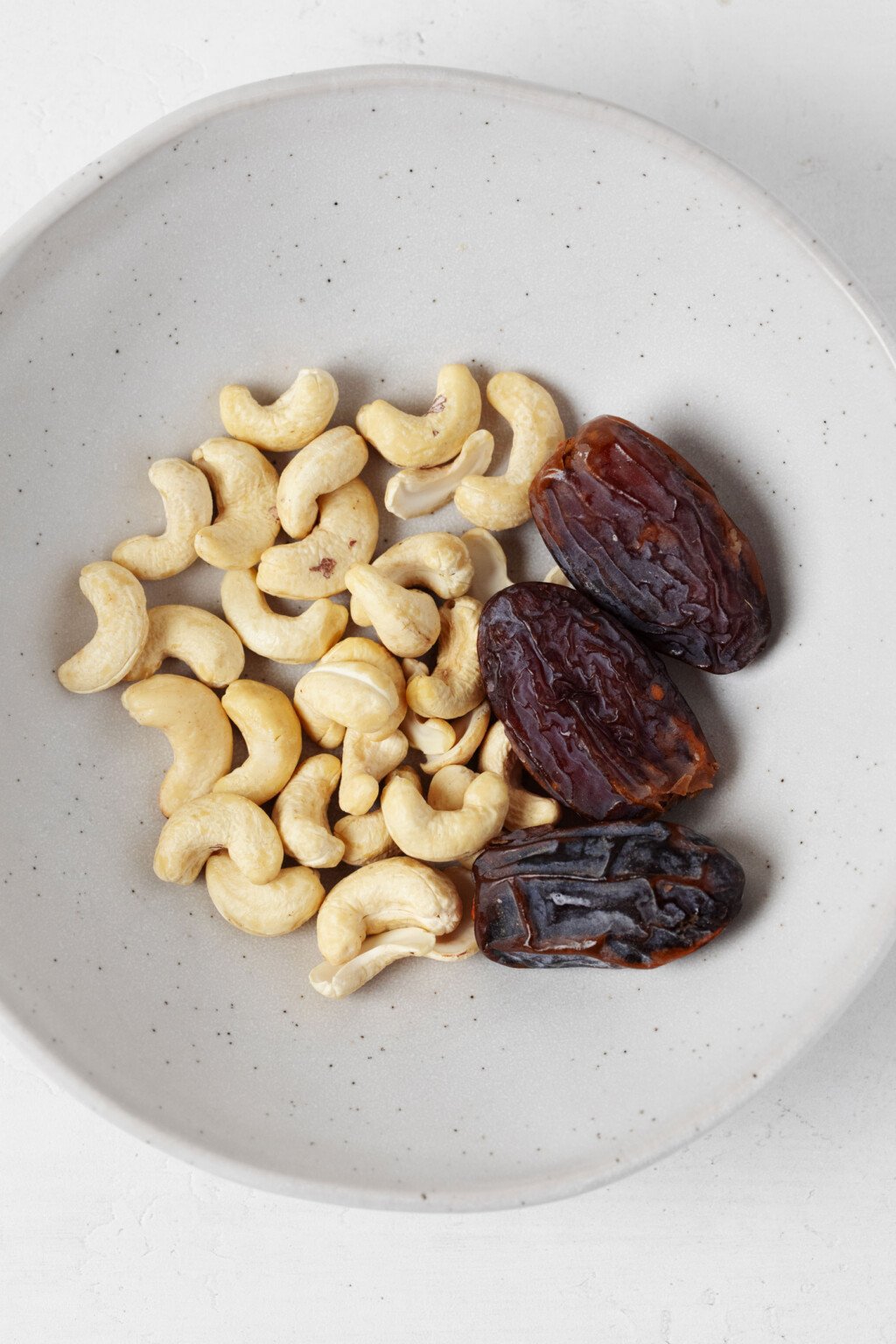 Cashews and pitted dates are pictured in a small, white ceramic bowl.