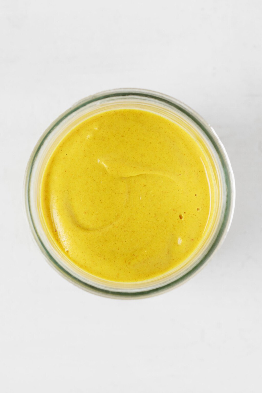 A clear glass jar is filled with a yellow-colored vegan honey mustard. The honey mustard has a creamy texture.