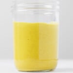 A clear glass jar is filled with a yellow-colored vegan honey mustard. The honey mustard has a creamy texture.