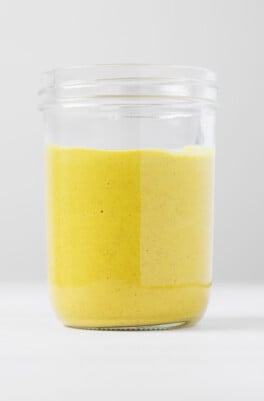 A clear glass jar is filled with a yellow-colored vegan honey mustard. The honey mustard has a creamy texture.