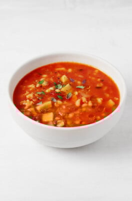 A bowl of root vegetable barley soup, made with a brothy tomato base, is served in a round white bowl. The bowl rests on a white surface.