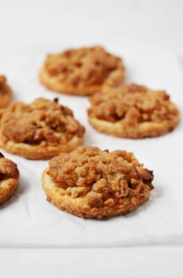 Plant-based apple pie crumble cookies are a deep golden brown, with buttery crumb topping. They've been laid on a white parchment surface.