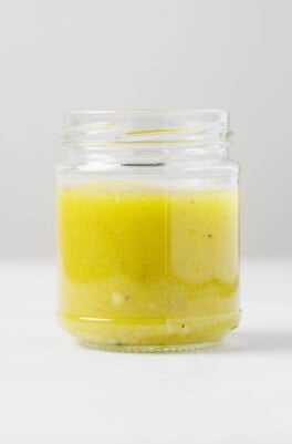 An image of a glass bottle, which is filled with a pale-yellow apple cider vinaigrette dressing. It rests on a white surface.