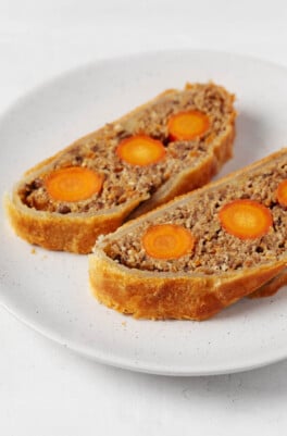 A vegan Wellington has been prepared with carrots and lentils at the center. Two slices of the puff-pastry-wrapped Wellington are arranged on a small, round white plate.