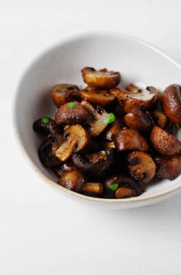 An asymmetrical white bowl is holding deep brown, air fryer mushrooms. The mushrooms are dotted with bright green herbs.
