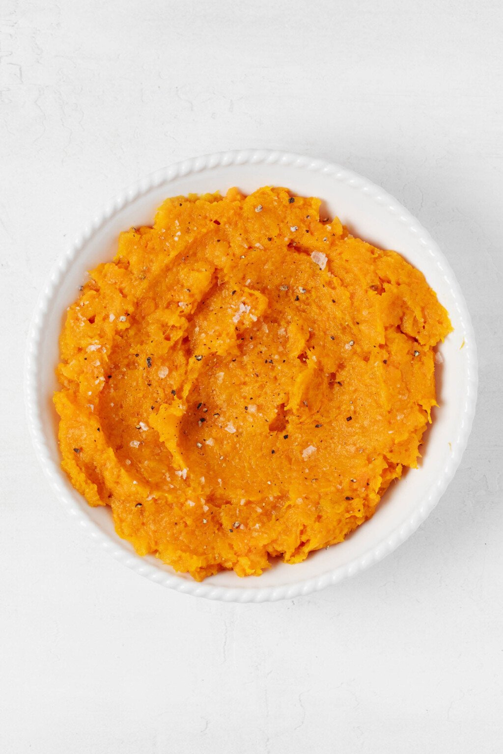 A white, round bowl holds a bright orange mixture of mashed sweet potato and butternut squash. 