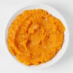 A white, round bowl holds a bright orange mixture of mashed sweet potato and butternut squash.