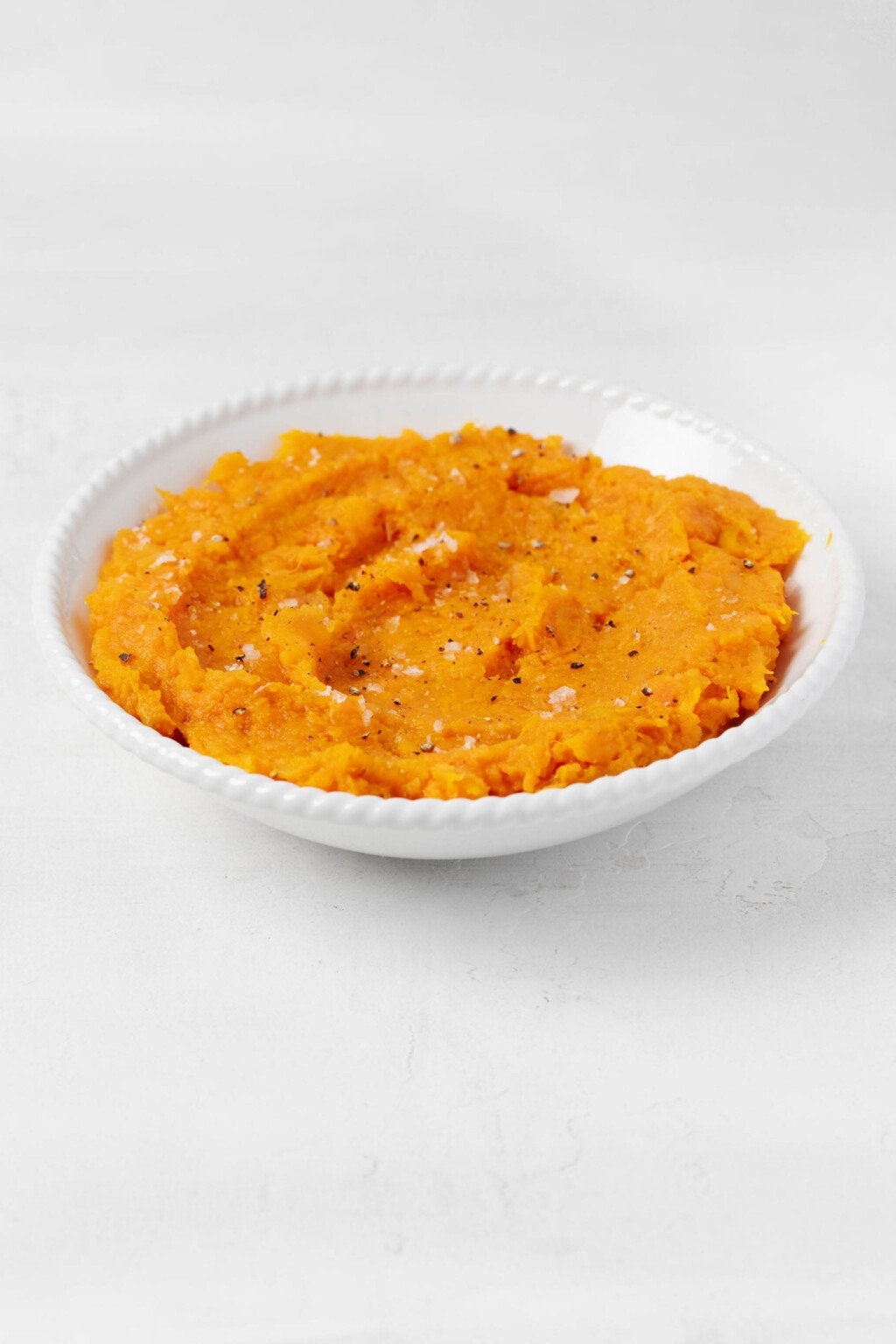 A white, round bowl holds a bright orange mixture of mashed sweet potato and butternut squash.