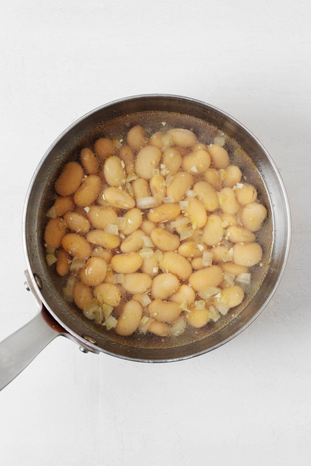 Beans, along with garlic and shallot and broth, are pictured in a silver saucepan.