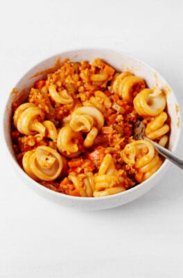 A round, white ceramic bowl rests on a white surface. It holds curly pasta shapes with a vegan bolognese sauce.