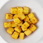A round, wide-rimmed white bowl holds bright golden baked curried tofu cubes.