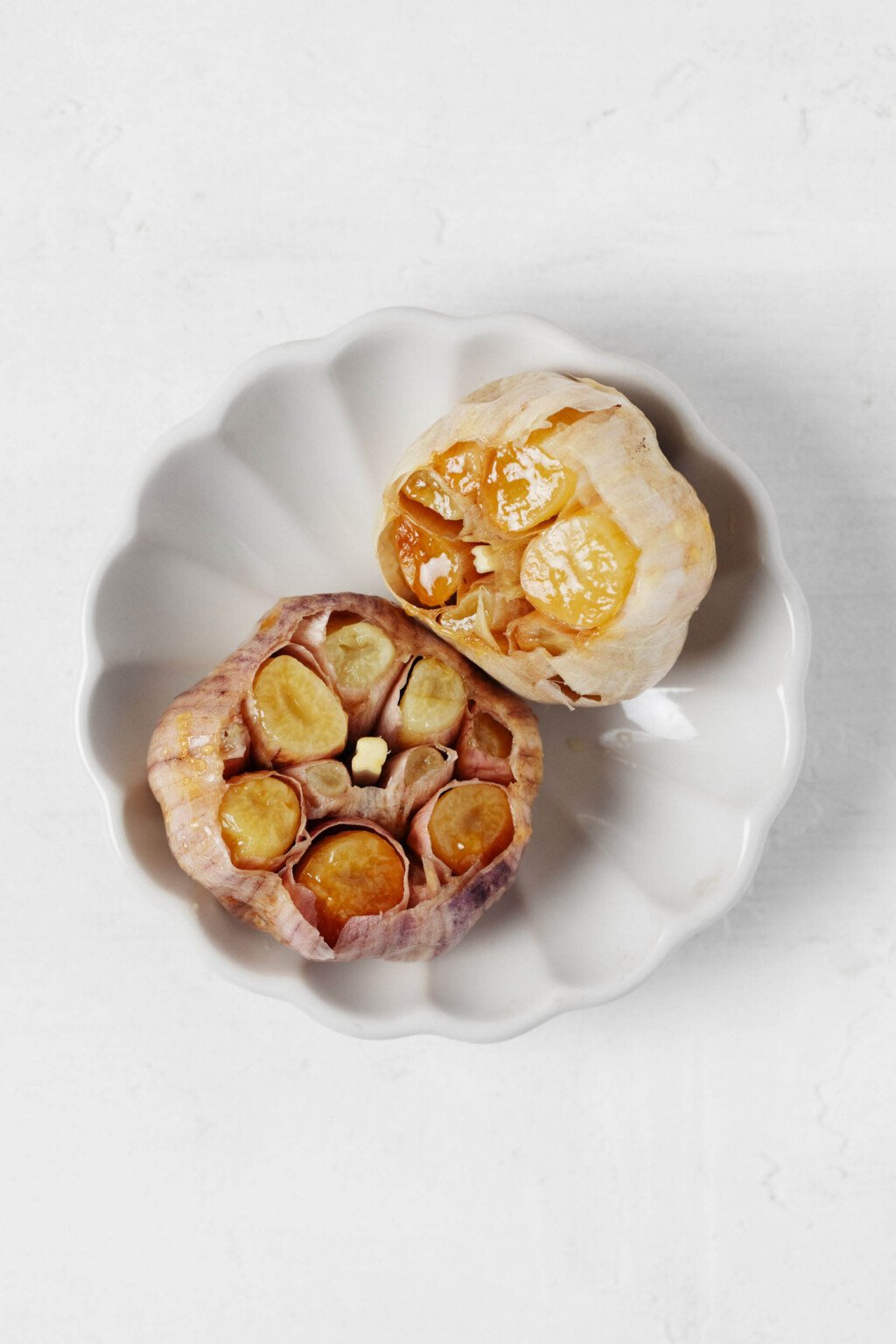Two bulbs of freshly roasted garlic are cut to expose caramelized cloves within. They rest on a small fluted plate.
