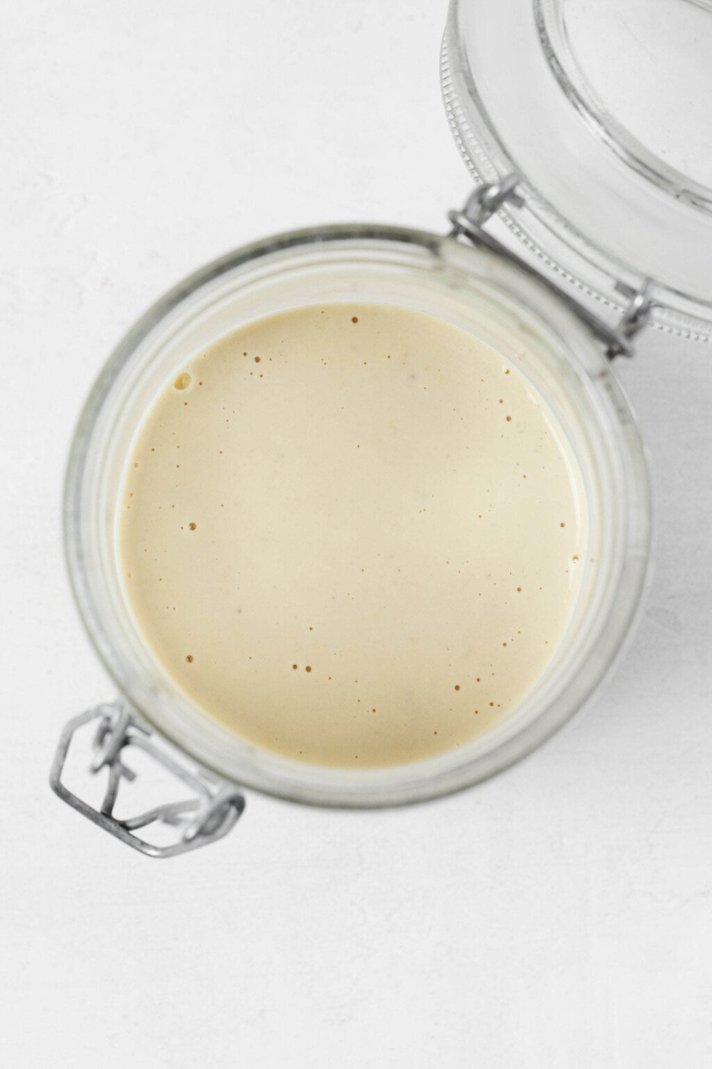 A jar of creamy white sauce is poured into a glass storage container. It rests on a white surface.