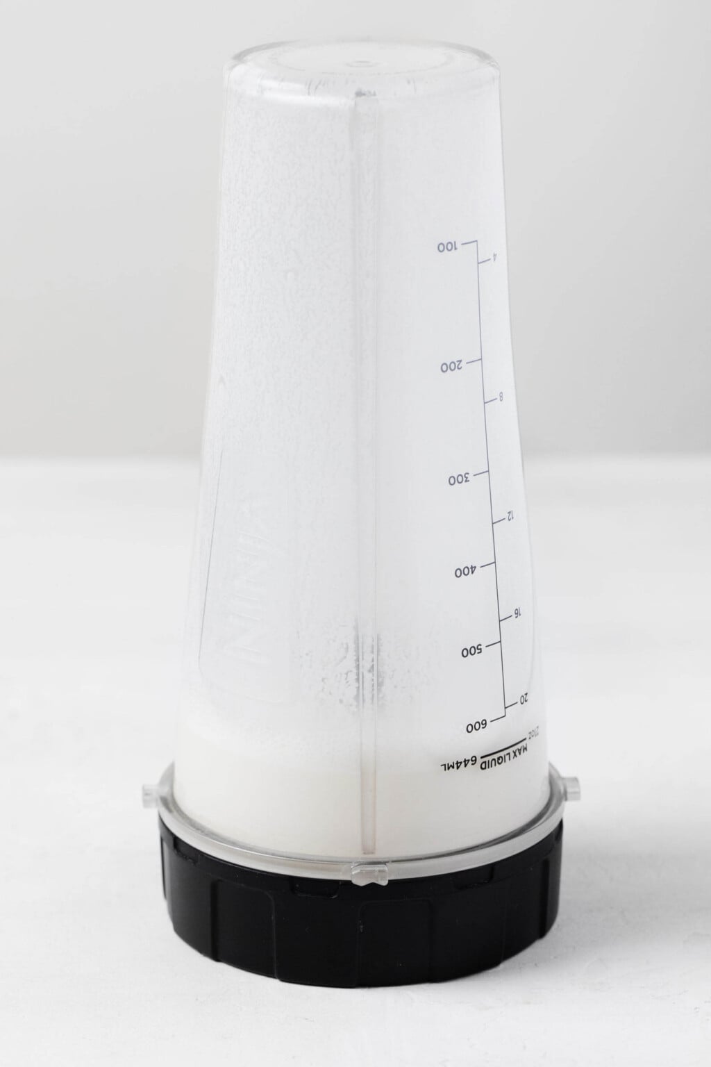 A small, personal blender is filled with a cup of pale colored cashew cream.